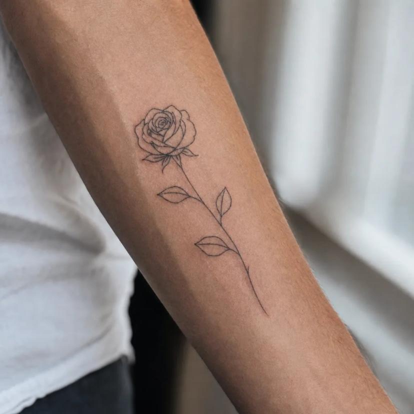 Tatouage rose fine-line minimaliste avec tige et feuilles sur l'avant-bras
